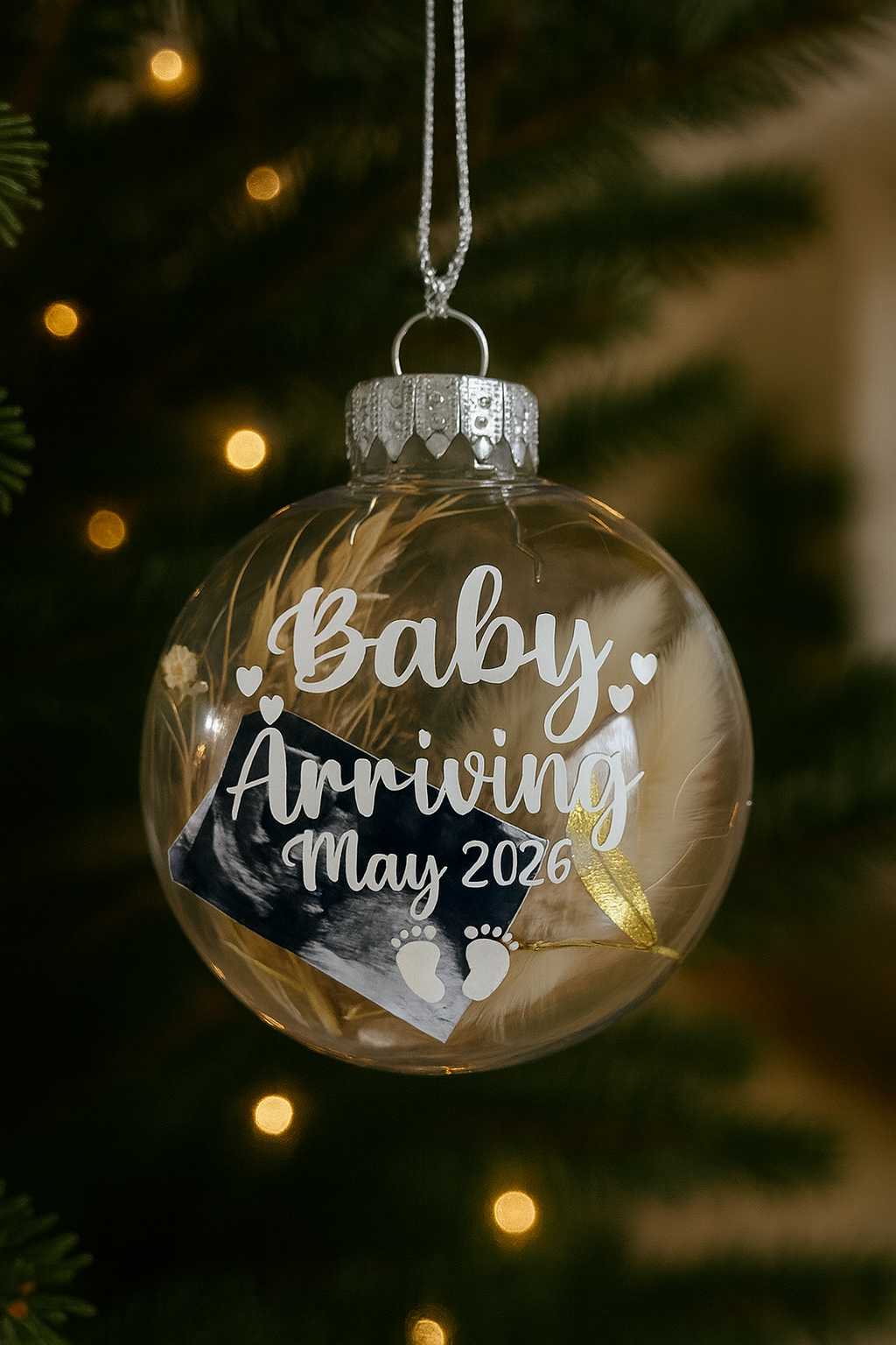 Clear Christmas ornament with 'Baby Arriving May 2026' text, hanging on a tree.