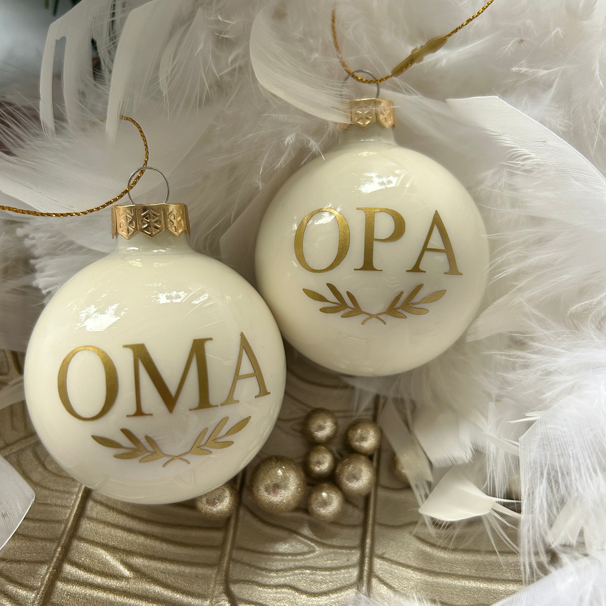 Two round glass ornaments with the letters 'OMA' and 'Opa' in gold vinyl, placed on a white surface with feathers and pearls around them.