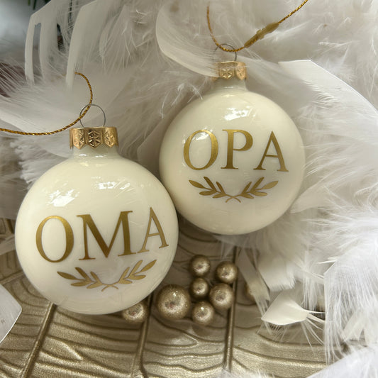 Two round glass ornaments with the letters 'OMA' and 'Opa' in gold vinyl, placed on a white surface with feathers and pearls around them.