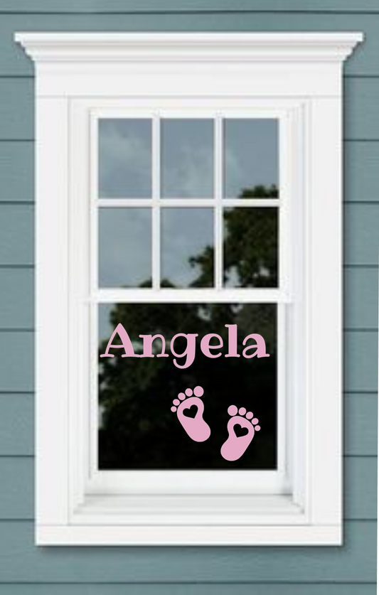 A window sticker with the name 'Angela' and a pair of baby feet on it, placed on a window sill.